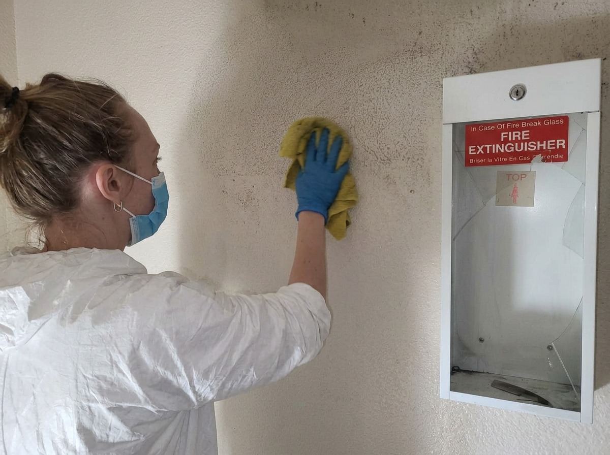 Fortis Restore technician in protective coveralls wiping smoke and soot residue from a wall beside a fire extinguisher cabinet